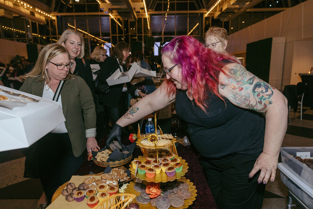 Milk Bank & Cookies on a table while people look through, woman in black with pink hair arranging cookies