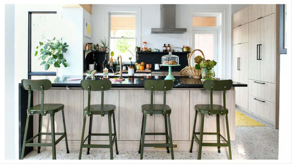 A bright, modern kitchen featuring a light wood cabinet island with olive green bar stools, black marble countertops and backsplash, integrated appliances, and a stainless steel hood above the range.