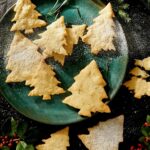 A dark green plate filled with Christmas tree-shaped shortbread cookies, some dusted with powdered sugar.
