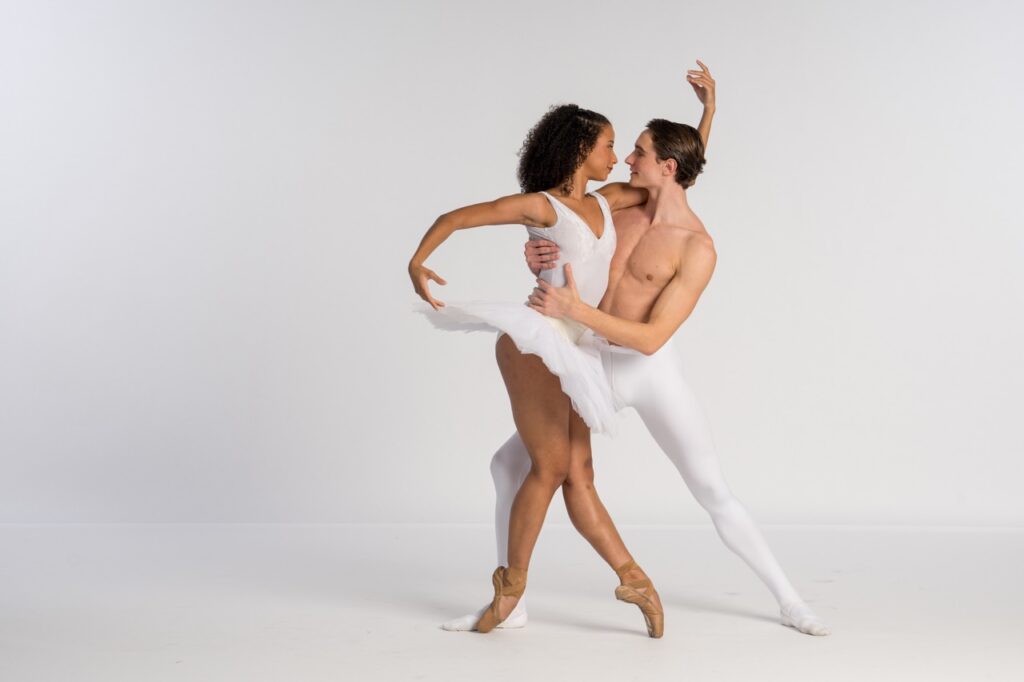 A male ballet dancer in white tights holds the waist of a female ballet dancer in a white dress.