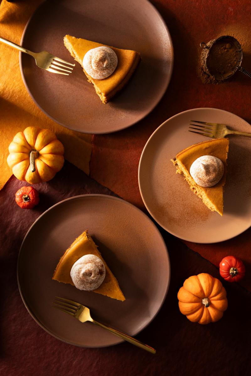 Three slices of pumpkin cheesecake on brown plates are arranged on a multicolored surface, each slice topped with a dollop of whipped cream and a sprinkle of cinnamon.