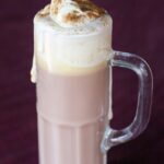 A tall glass mug of hot chocolate with melted whipped cream on top, dusted with cinnamon, set against a dark purple background.