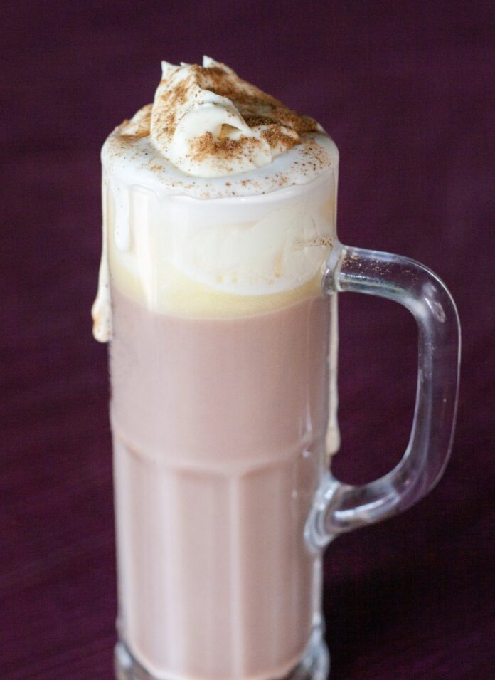 Raelynn Gigler of Dish Osteria Heats Up with Hot Chocolate TABLE Magazine A tall glass mug of hot chocolate with melted whipped cream on top, dusted with cinnamon, set against a dark purple background.