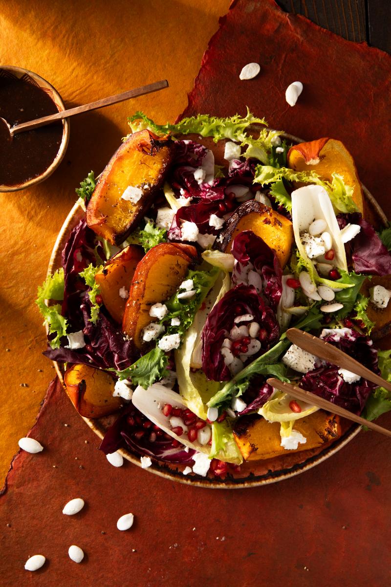 A colorful roasted pumpkin salad is presented on a plate with red and green lettuce, goat cheese, and pomegranate seeds.