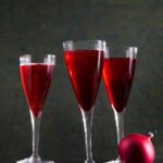 Three stemmed champagne flutes filled with a red Sand Hill Kir Royale cocktail and a single red holiday ornament.