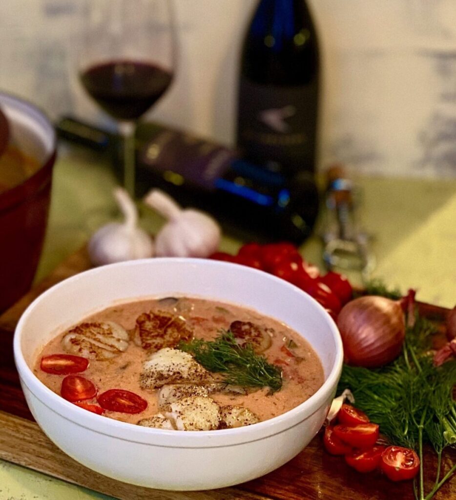 A white bowl of Seafood Chowder sits beside a glass of red wine, its bottle, garlic, onions, and tomatoes.
