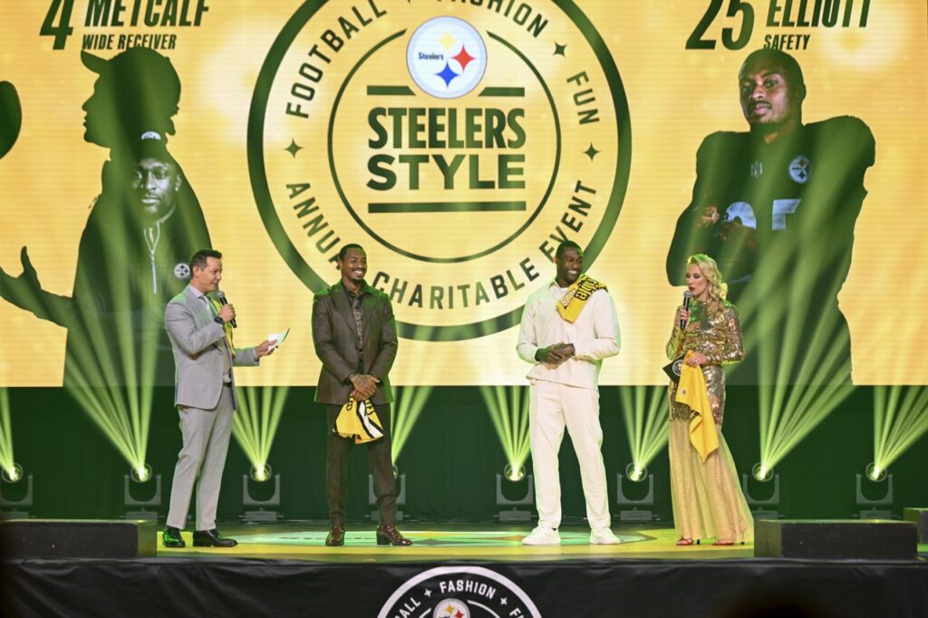 Three men in light grey, dark grey, and white suits stand on a stage beside a woman in a yellow sequins dress.