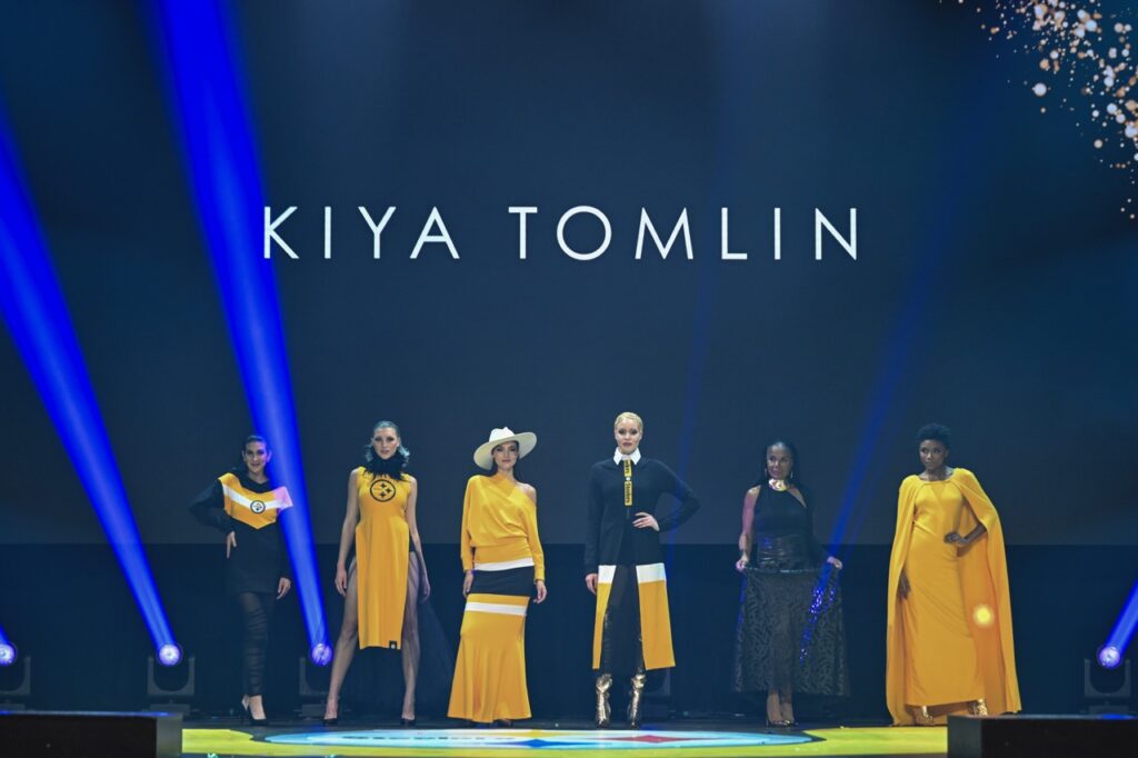 Six women wear various black and gold dresses on a stage.
