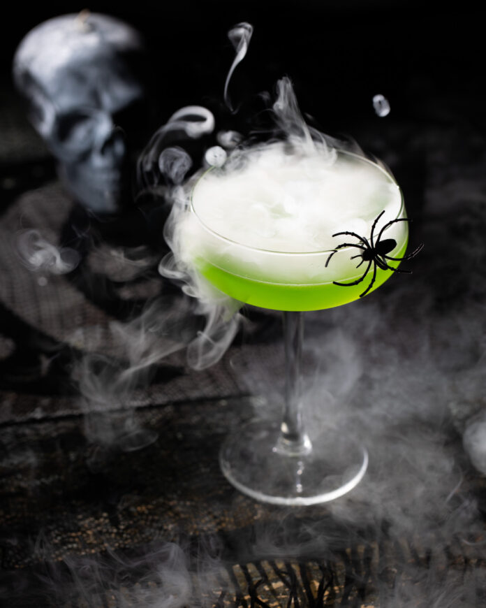 TABLE Magazine Halloween Cocktail Witch's Brew A green cocktail on a black background, styled for Halloween with spiders and smoke surrounding it