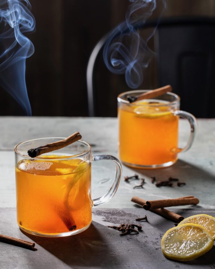 Two glasses hold a hot toddy orange liquid with lemon slices inside and a smoking cinnamon stick on top to represent Teddy Swims in a cocktail.