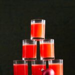 A tall pyramid of small glasses filled with a vibrant red-orange "The Babysitter" cocktail, with a small red Christmas ornament next to it.