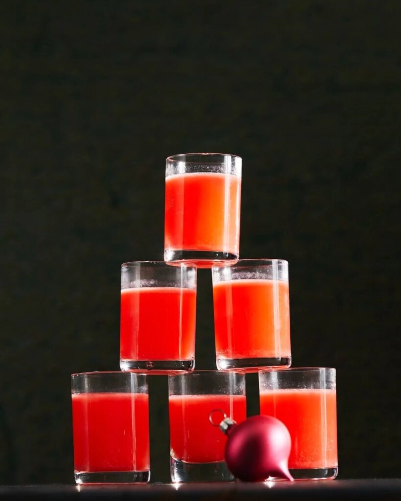 A tall pyramid of small glasses filled with a vibrant red-orange "The Babysitter" cocktail, with a small red Christmas ornament next to it.