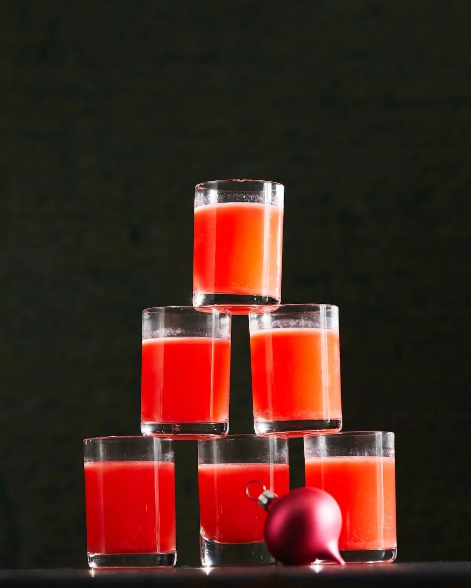 A tall pyramid of small glasses filled with a vibrant red-orange "The Babysitter" cocktail, with a small red Christmas ornament next to it.
