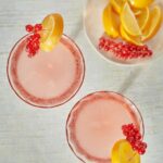 Two pink Fitzgerald gin cocktails garnished with red currants and lemon slices on a white wooden surface.