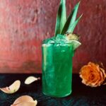 A vibrant aqua green cocktail in a tall glass with pineapple leaves, a pineapple wedge and lime wedge as garnish with an orange rose and rose petals in the foreground.