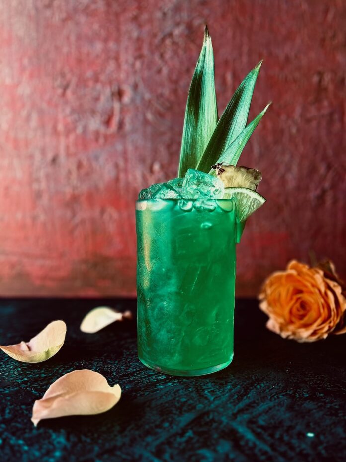 The Life of a Showgirl Cocktail TABLE Magazine A vibrant aqua green cocktail in a tall glass with pineapple leaves, a pineapple wedge and lime wedge as garnish with an orange rose and rose petals in the foreground.