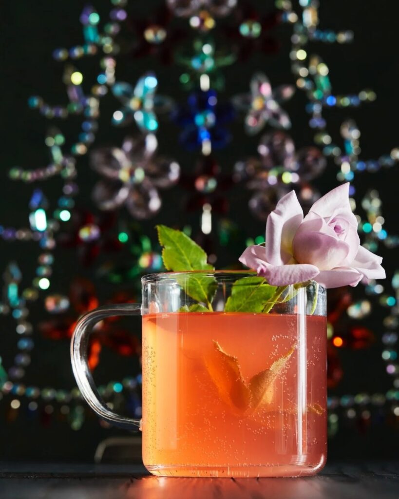 A glass mug of peacekeeper cocktail, garnished with a rose and a lemon peel, sits in front of a blurry, colorful background.
