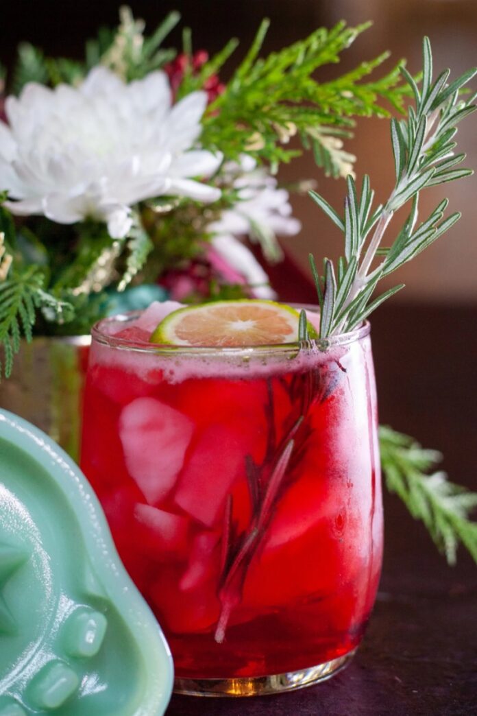Yule Margarita TABLE Magazine A red Yule Margarita cocktail on ice, garnished with a lime slice and a rosemary sprig, with holiday decor in the background.