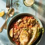 A blue bowl holdsbraised cabbage and beans topped with a lemon slice and accompanied by a fork, spoon, and glass of lemon water.