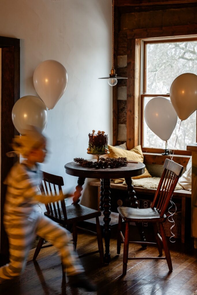 A blurred image of a child in striped pajamas running past a small wooden table holding a moss and chocolate birthday cake, surrounded by white balloons and a window with a snowy view.