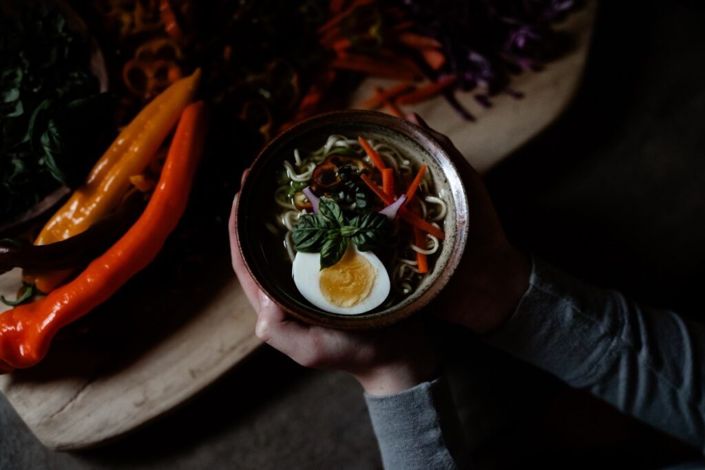 A hand holding a bowl of ramen soup topped with half a hard-boiled egg, fresh basil, shredded carrots, and dark vegetables, with colorful vegetables like orange peppers in the background.