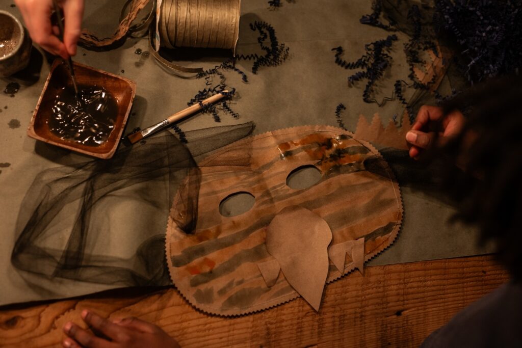 A child's hands painting and gluing a brown paper "Wild Thing" mask on a dark surface, with crafting supplies like ribbon and a cup of brown paint nearby.