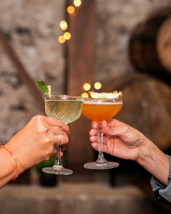 A Community of Purpose TABLE Magazine Two people clinking cocktail glasses, one clear and one orange, in a toast at a non-profit mixer or community event, with bokeh lights and wine barrels in the background.