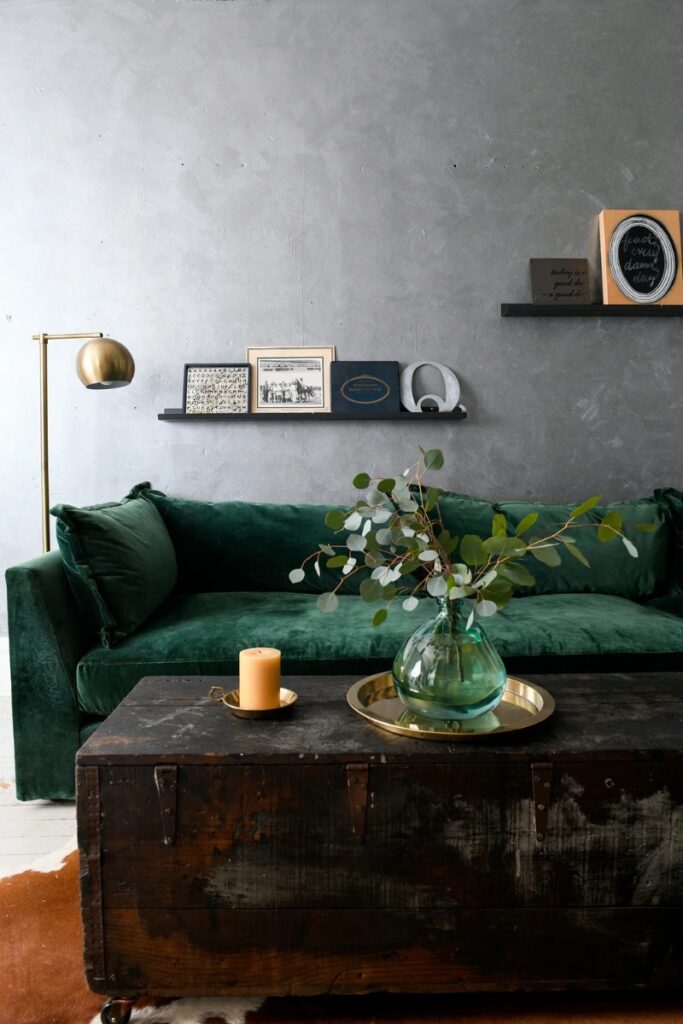 A deep emerald green velvet sofa sits against a textured gray wall, accented by a gold floor lamp and a dark antique wooden trunk used as a coffee table with a glass vase of eucalyptus and a candle on a brass tray.
