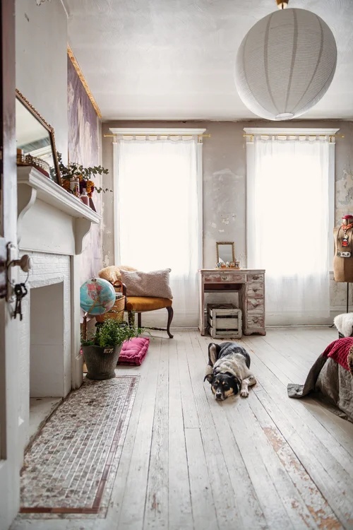 A dog lying on distressed white wood floors in a minimalist, vintage-style room with two bright windows, a white fireplace, an antique wooden desk, and a large white paper globe lantern.