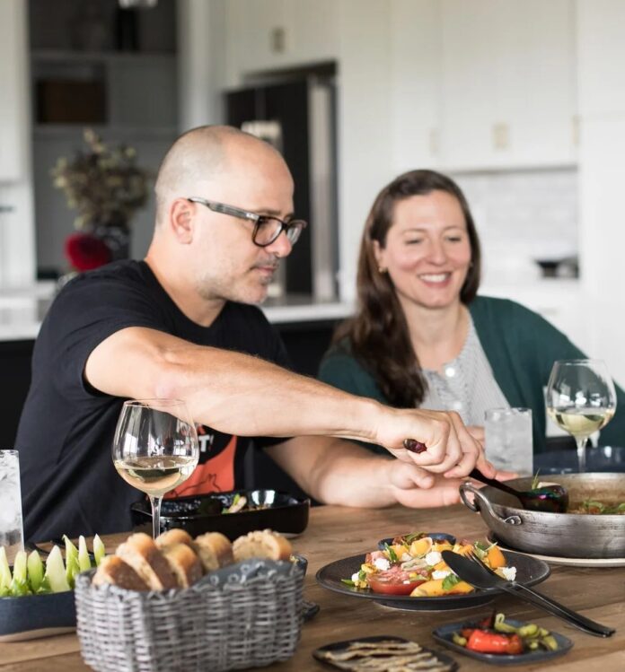 Chef Justin Severino, wearing glasses and a black shirt, sits at a wooden table with his wife, Hilary Prescott Severino, smiling in the background, as they share a family-style meal featuring wine, bread, and a colorful tomato and peach salad.