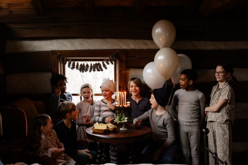 A group of children in pajamas gathered around a small wooden table with a moss-and-bark birthday cake and lit candles, with white balloons floating nearby in a rustic log cabin.