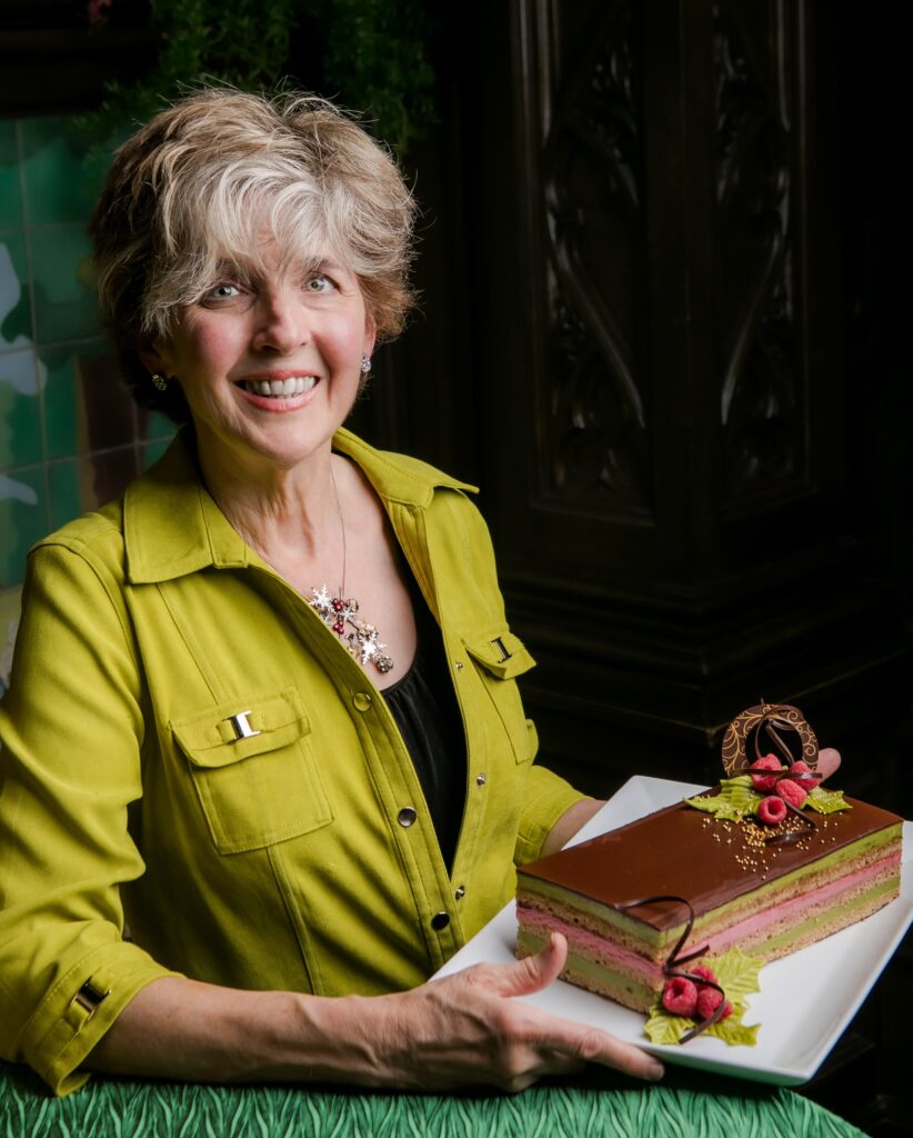 A women in a green button down shirt holding a festive opera torte, layered in pink and green with chocolate on top, on a white plate decorated with raspberries, curls of chocolate, green leaves and gold leaf.