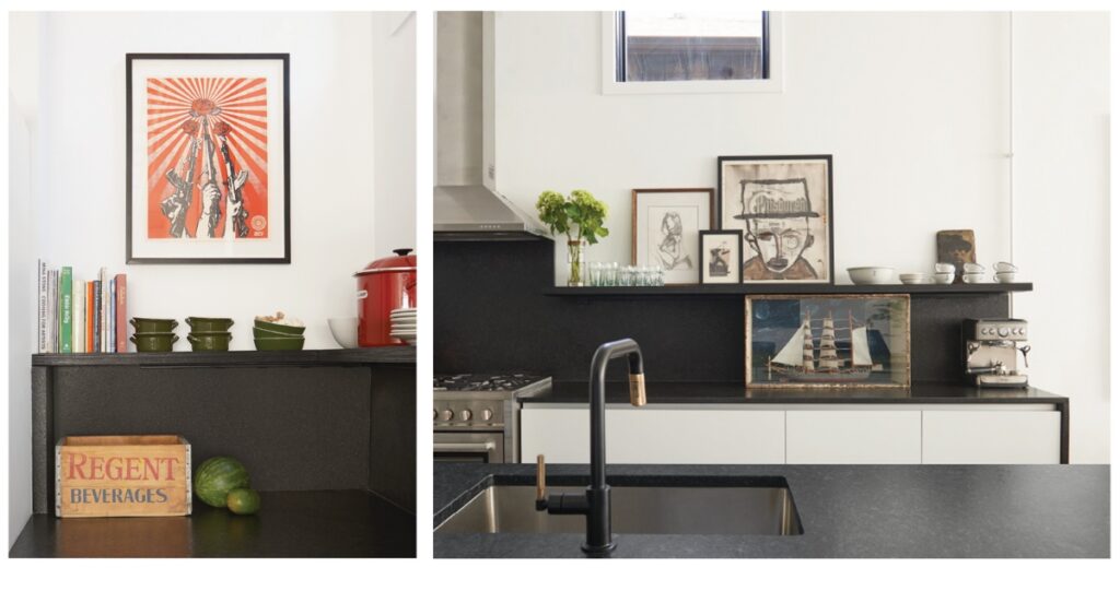 A modern kitchen counter with a black sink and faucet, featuring a curated display of framed art including a ship painting, small bowls, a toaster, and fresh greenery on a black floating shelf against a white wall.