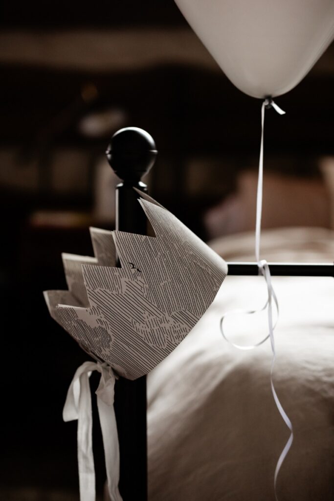 A paper crown, tied with a white ribbon, and a white balloon hanging from a black metal bedpost, suggesting a minimalist birthday celebration theme.