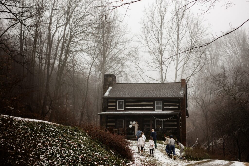 A rustic log cabin in a foggy, snow-dusted, bare winter forest with a group of children in white and neutral clothing walking toward it.