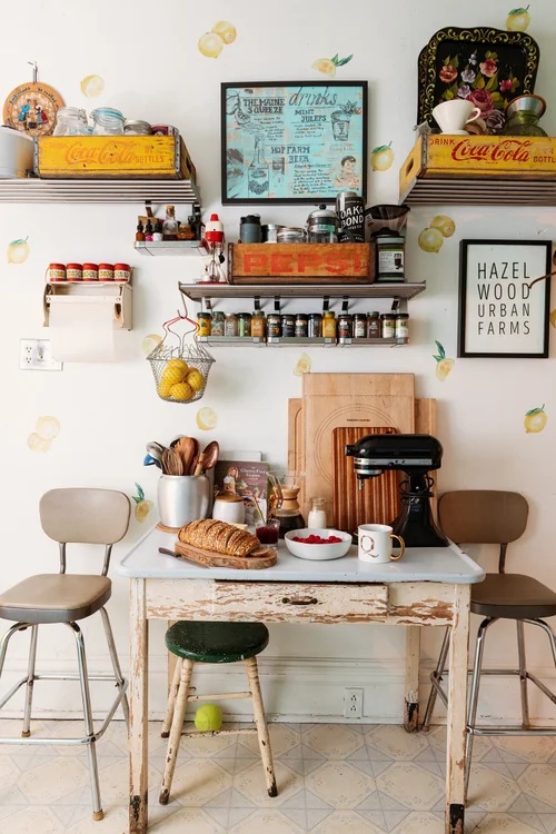 A vintage kitchen corner with white walls decorated with lemon decals and framed retro art, featuring metal shelving with Coca-Cola crates, spice racks, a small dining table with a bread loaf, and two bar stools.