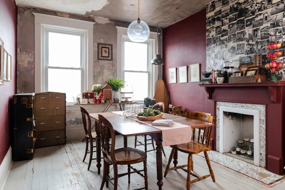 A vintage-style dining room with distressed white wooden floors, burgundy walls, a collage photo wall, and a wooden table with mismatched chairs, showcasing a maximalist home interior.