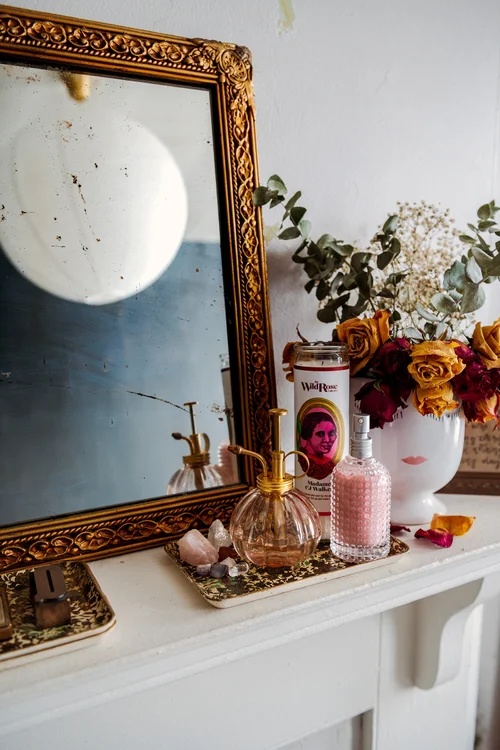 A vintage white fireplace mantle decorated with an ornate gold framed mirror, dried roses and eucalyptus in a face-shaped vase, and various pink and gold glass perfume bottles and personal items.