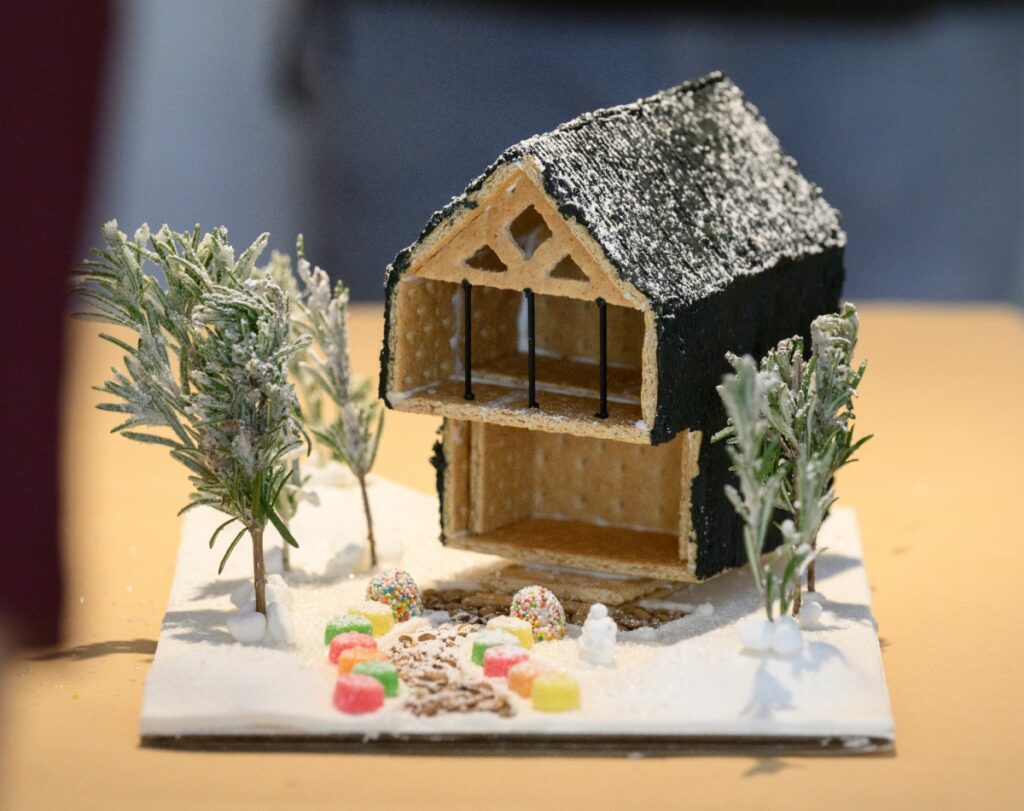 A winning modern gingerbread house design made with graham crackers and black cladding, featuring a frosted rosemary Christmas tree and gumdrop candy garden on a snowy white icing base.