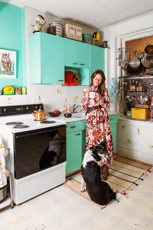 A woman wearing a floral dress stands in a vintage kitchen with bright mint green cabinets and lemon decals on the wall, holding a mug while a black and white dog sits at her feet.