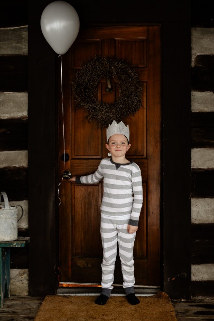A young boy in gray and white striped pajamas and a paper crown stands on a porch holding a gray balloon, with a wreath on the dark wooden log cabin door behind him.