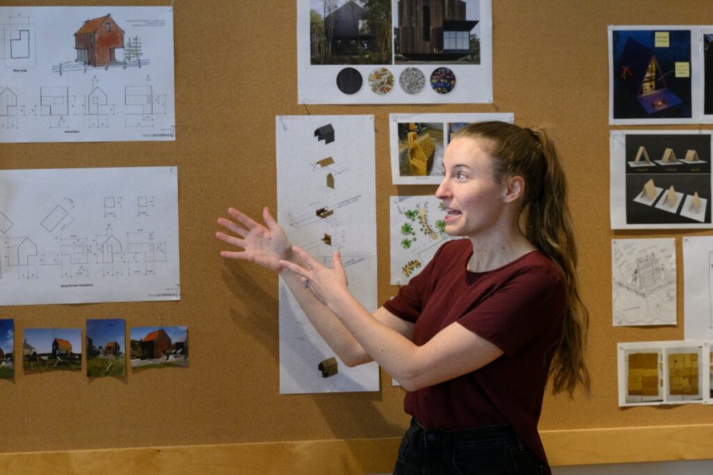 A young woman giving a presentation with her hands, standing in front of a cork board displaying architectural drawings, photographs, and design concepts.