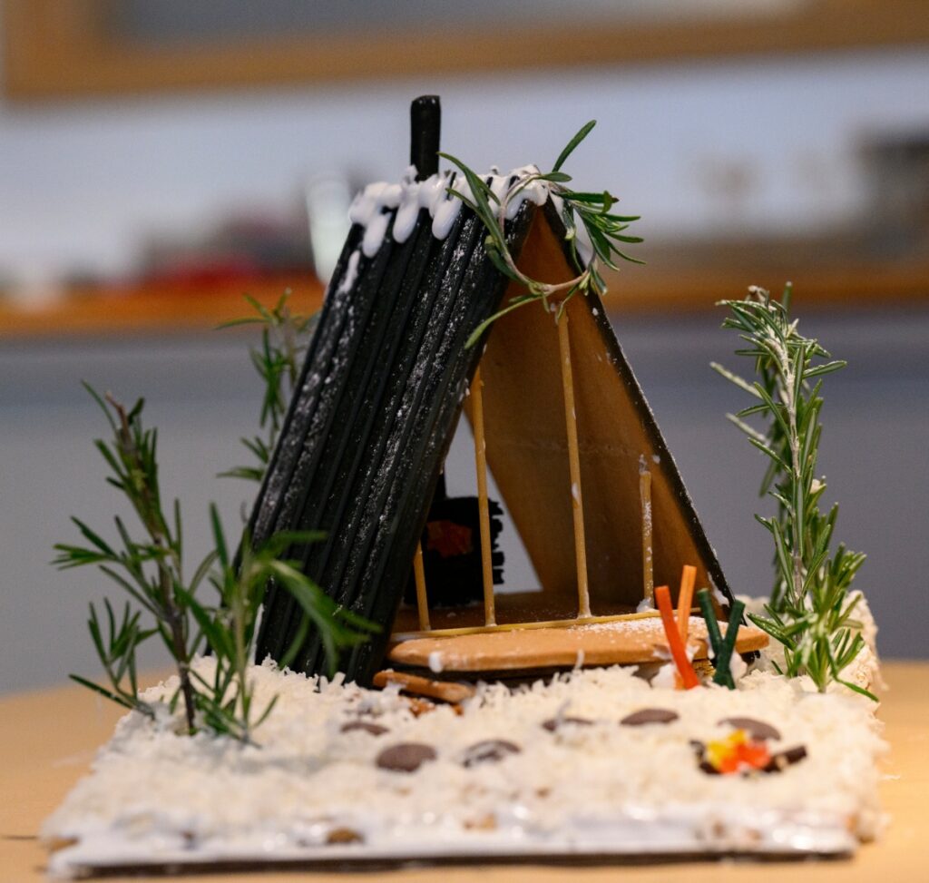 A modern A-frame gingerbread ski chalet, or 'Slope Haus,' with a black roof made of pocky sticks and snow-like icing, surrounded by rosemary trees in a baking contest.