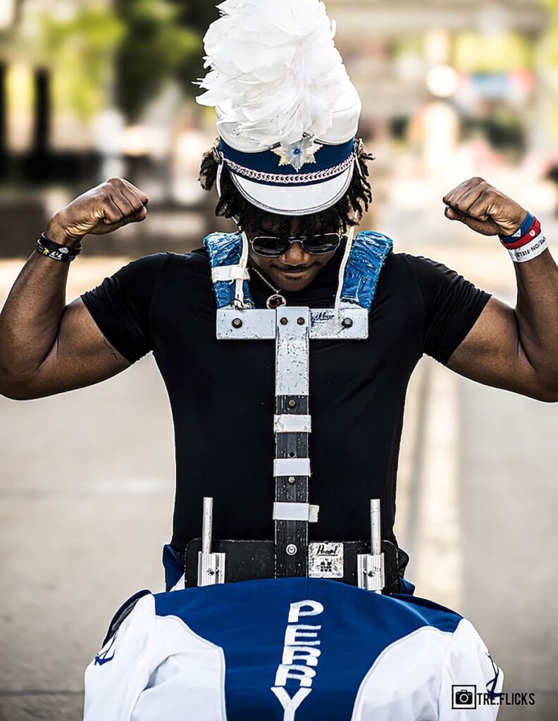 A man in a marching band hat carries a snare on his body.
