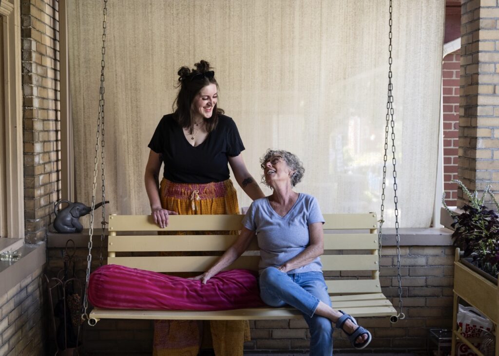 An older woman sits on a porch swing as a younger woman looks down at her from behind it.