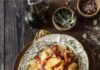Ampezzo-Style Potatoes A plate of Ampezzo-style potatoes (sliced potatoes) with crispy speck and red onions, topped with black pepper and served on a patterned rustic plate with silver spoons and bread.