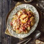 A plate of Ampezzo-style potatoes (sliced potatoes) with crispy speck and red onions, topped with black pepper and served on a patterned rustic plate with silver spoons and bread.