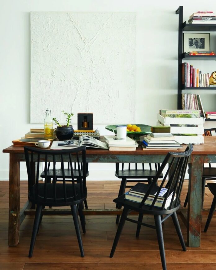 A modern dining area featuring a light wood rectangular table, black spindle chairs, and a black metal and glass shelving unit displaying woven baskets and decor, contrasting against white walls.