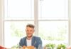 Chef Joey Hilty of The Vandal Builds a Fall Feast Chef Joey Hilty, smiling and wearing a denim shirt, sits at a dark table filled with a large family-style meal including a Caesar salad, plates of sliced meat with sauce, and glasses of red wine, set against a bright window.
