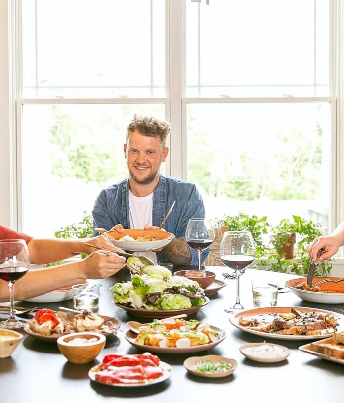 Chef Joey Hilty of The Vandal TABLE Magazine Chef Joey Hilty, smiling and wearing a denim shirt, sits at a dark table filled with a large family-style meal including a Caesar salad, plates of sliced meat with sauce, and glasses of red wine, set against a bright window.